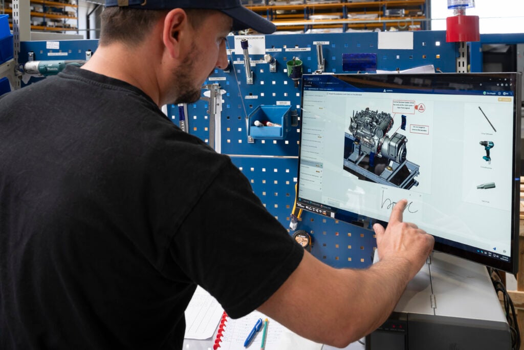 An operator at e-power uses a touchscreen to follow digital work instructions for assembling a power unit. The screen displays a detailed 3D image of an engine with annotations and tool references, powered by the Azumuta platform. In the background, tools are neatly organized on a blue pegboard, and a notebook and pens are visible on the workstation.