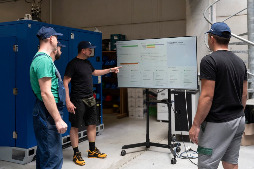 A team leader at e-power points to a large digital dashboard during a shop floor meeting. The screen displays quality metrics, audit scores, production progress, and internal updates. Several team members in work attire and caps are gathered around, engaging with the data being discussed.