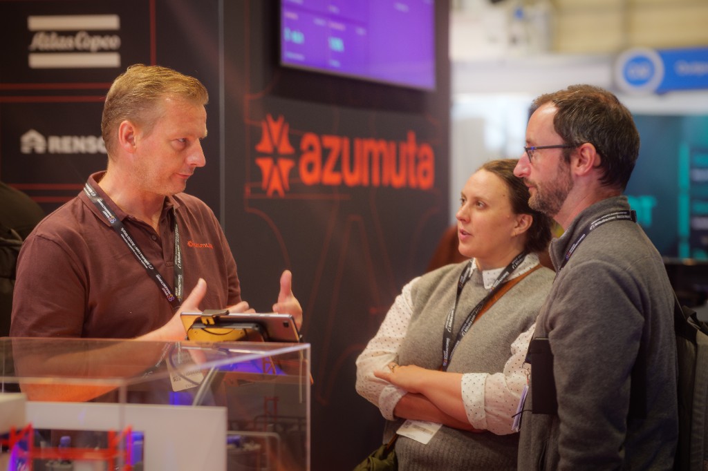 An Azumuta team member holding a tablet talks with two trade show visitors at the Azumuta booth, in front of a dark wall showing the Azumuta logo and customer logos including Atlas Copco and Renson.