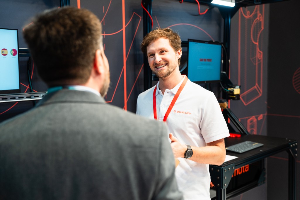 A smiling Azumuta team member in a white polo talks with a visitor at the Azumuta booth during Hannover Messe, with screens and shop-floor demo equipment in the background.