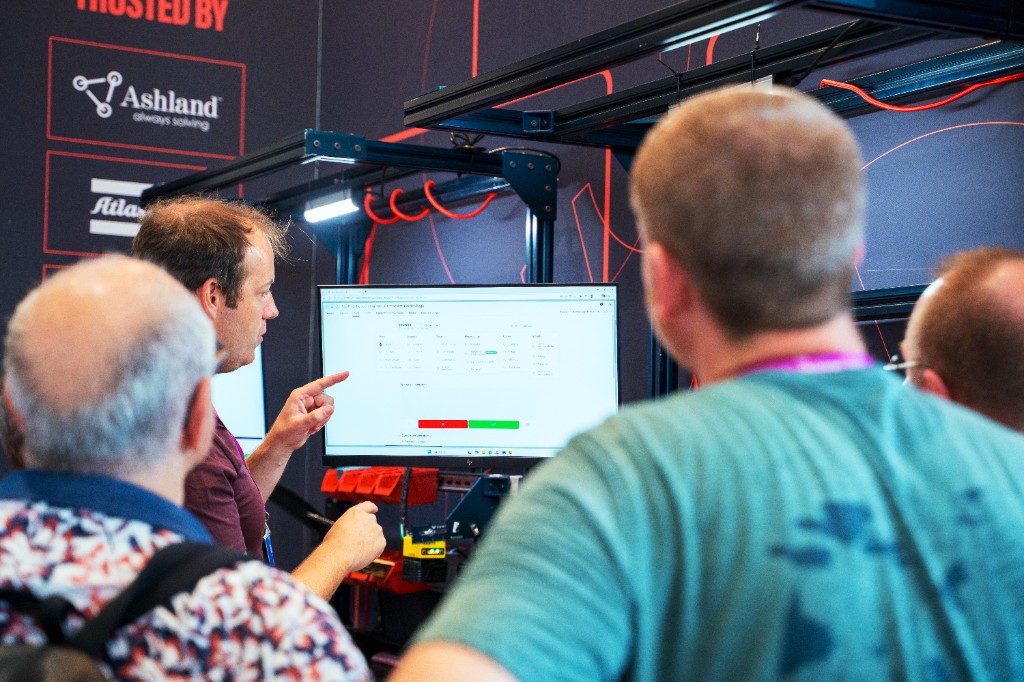 An Azumuta team member demonstrates the software on a large screen to a group of attentive visitors at the Tech For Industry trade show in Paris. The booth wall shows a 'Trusted by' section with customer logos including Ashland.